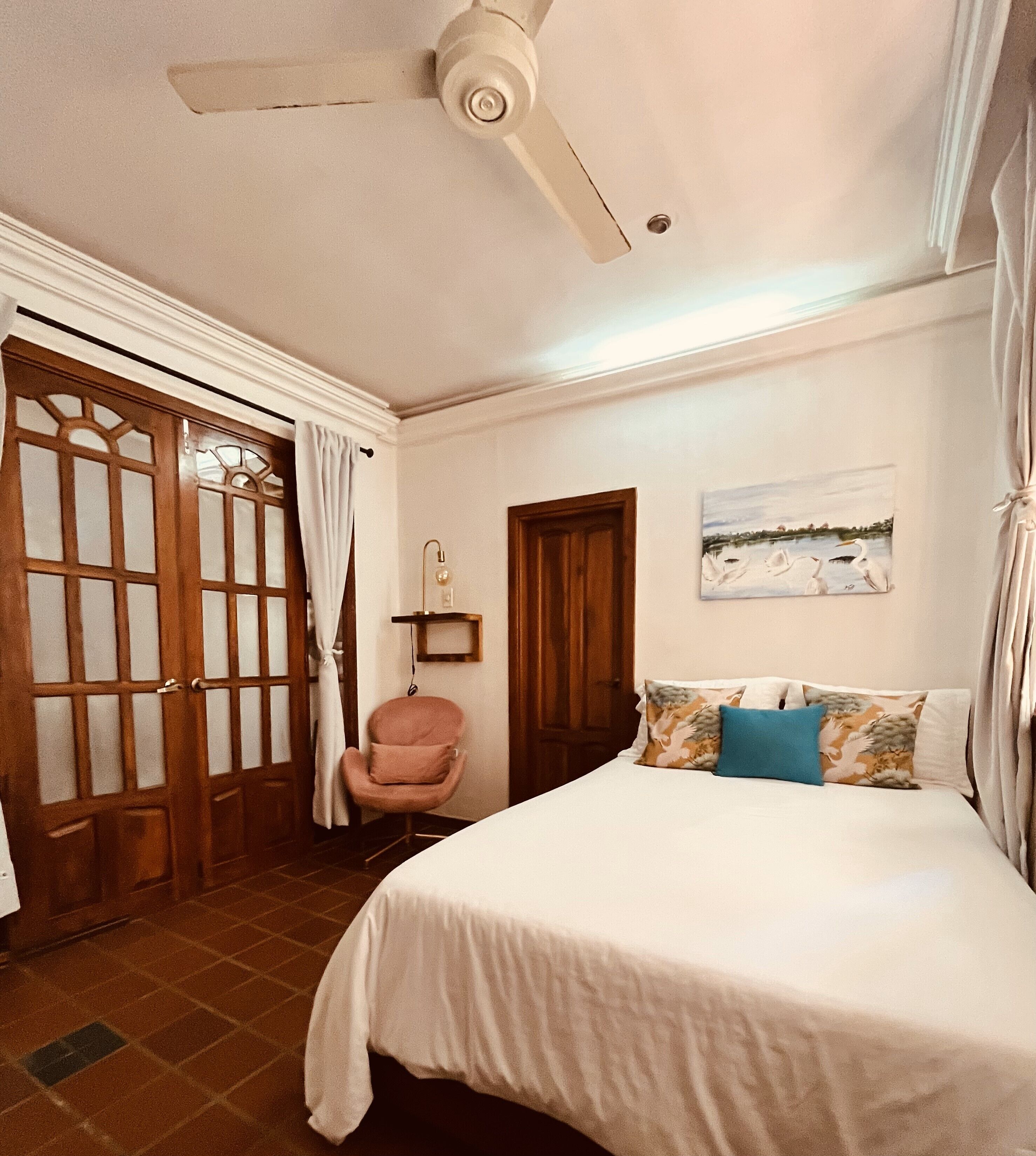 Un dormitorio con cama, una silla, ventilador de techo, una puerta y un cuadro en la pared.