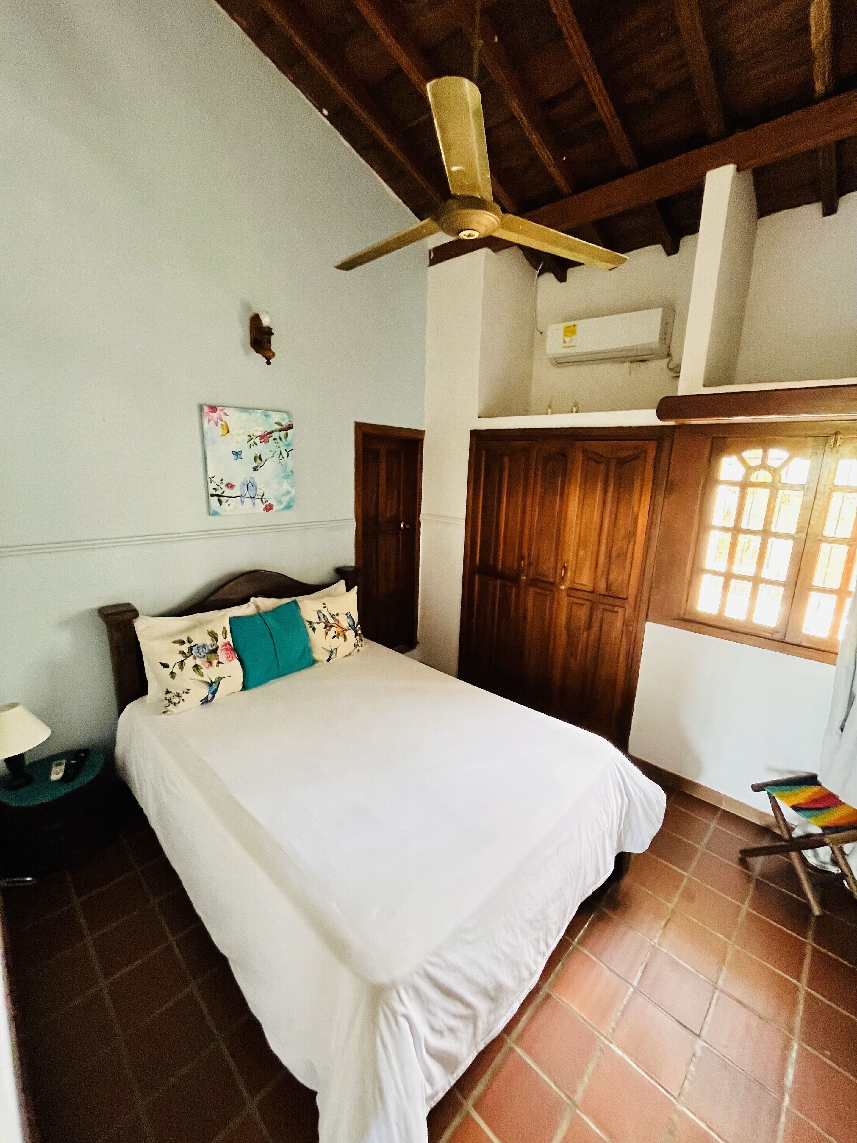 A bedroom with a bed, a ceiling fan, wooden wardrobe, and a window with sunlight.