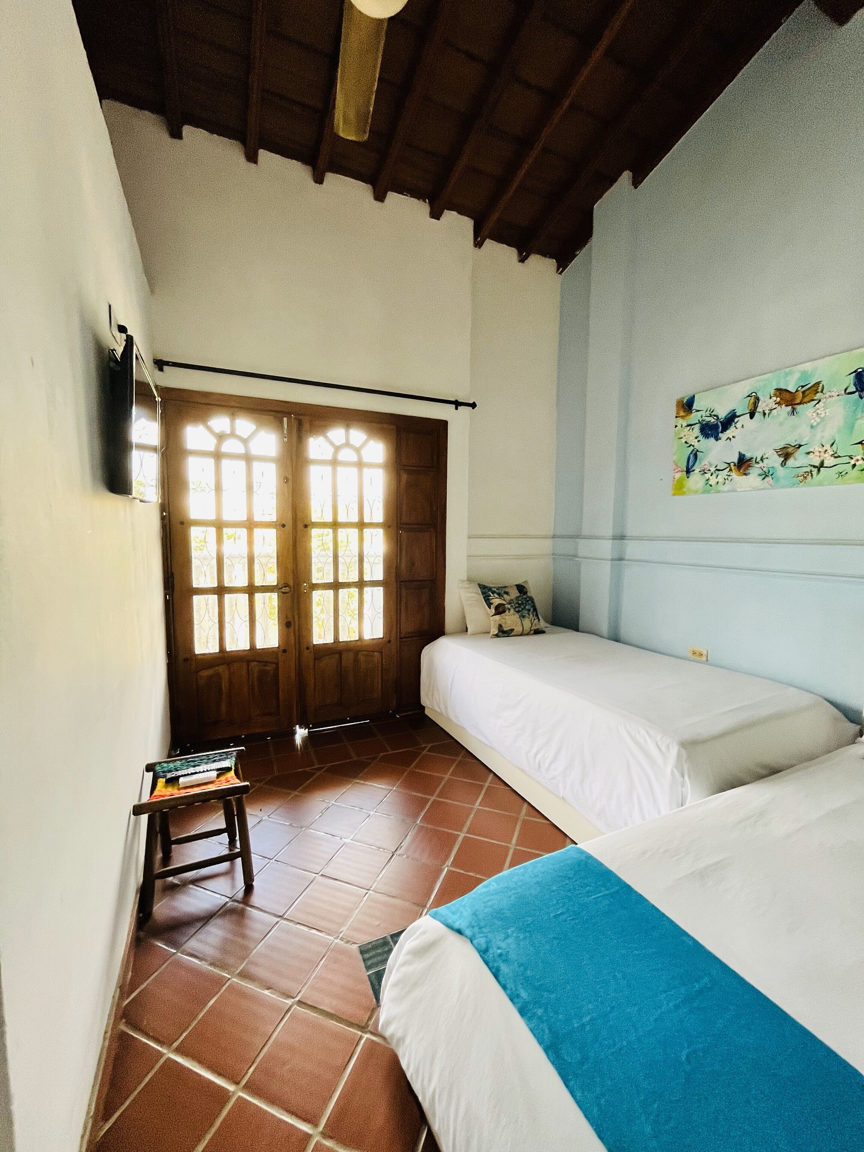 Una habitación con dos camas, una puerta de madera con paneles de vidrio, una mesita con una silla y un cuadro en la pared.