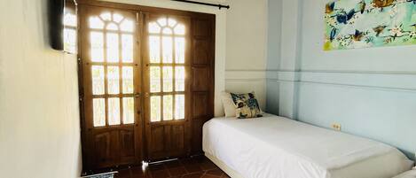 Una habitación con dos camas, una puerta de madera con paneles de vidrio, una mesita con una silla y un cuadro en la pared.