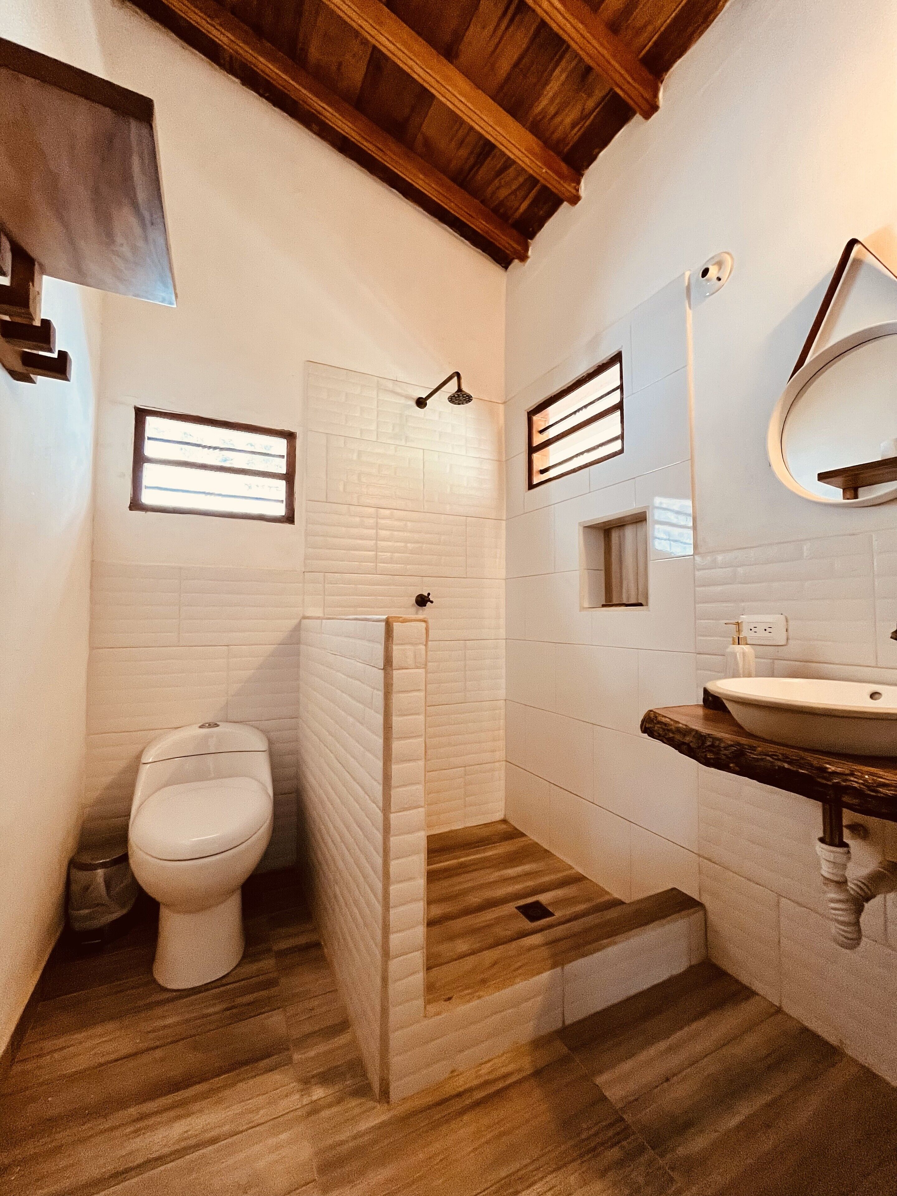 A bathroom with a wooden floor, white tiles, a toilet, a shower, and a sink with a wooden countertop.