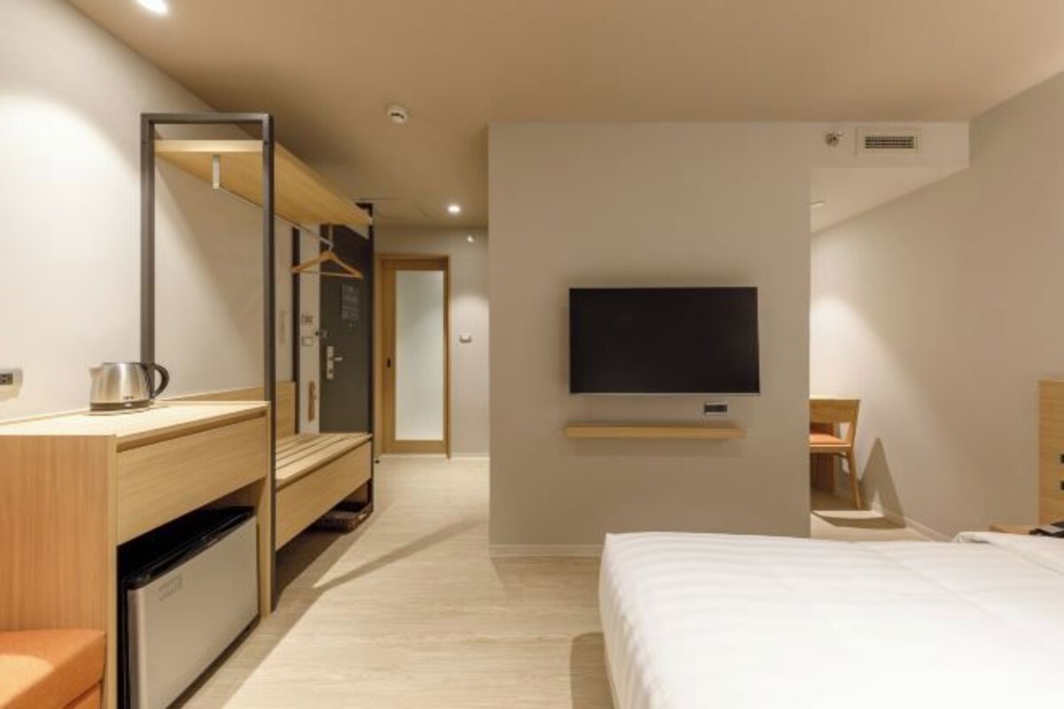 A modern hotel room with a bed, a TV mounted on the wall, a wooden dresser, a mirror, and a small kitchenette.