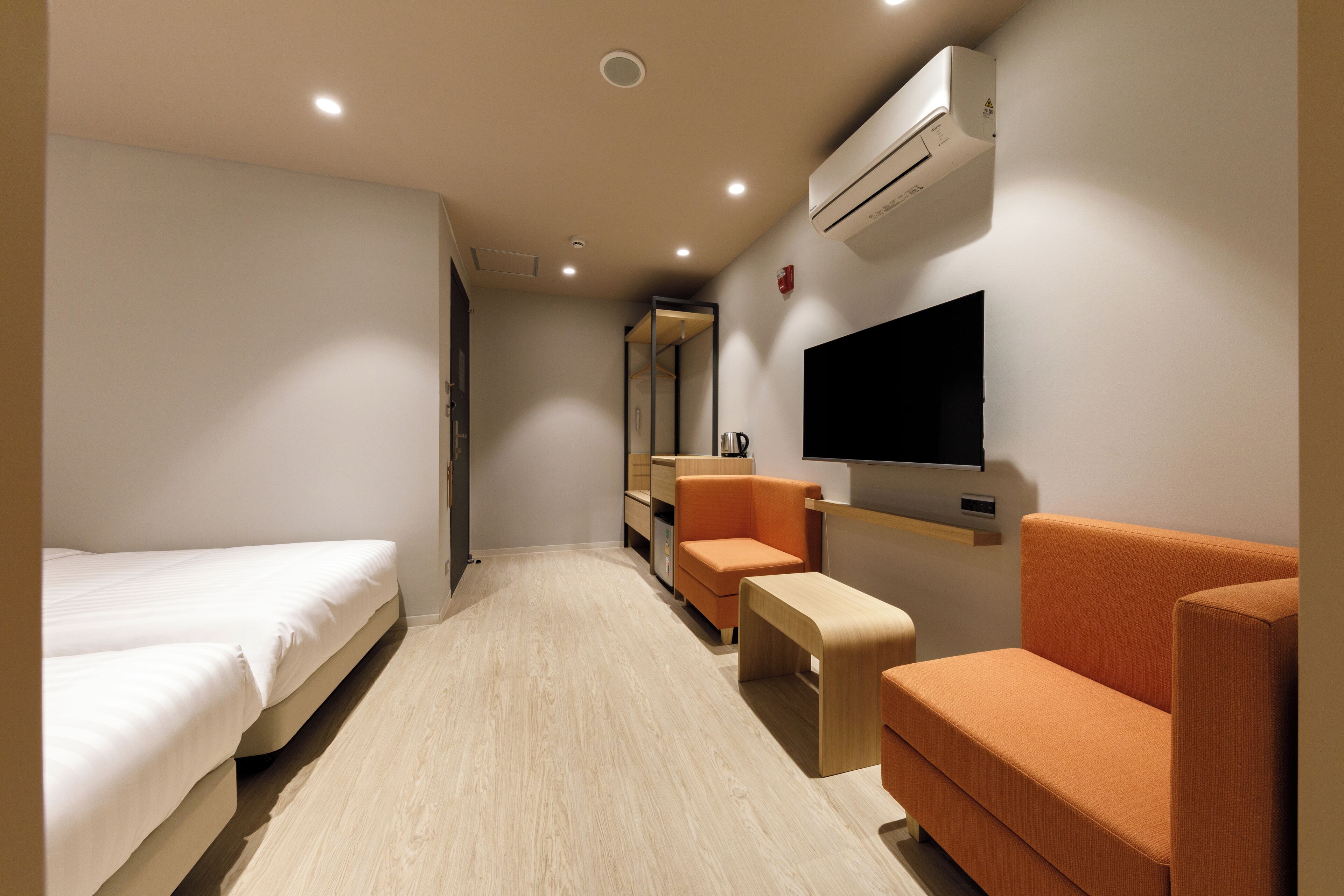 A hotel room with a bed, two orange armchairs, a small table, a TV, and an air conditioning unit.