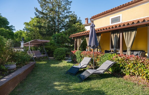 Ein Terrassenbereich mit zwei Liegestühlen, einem überdachten Sitzbereich und einem gelben Gebäude mit einem Ziegeldach.