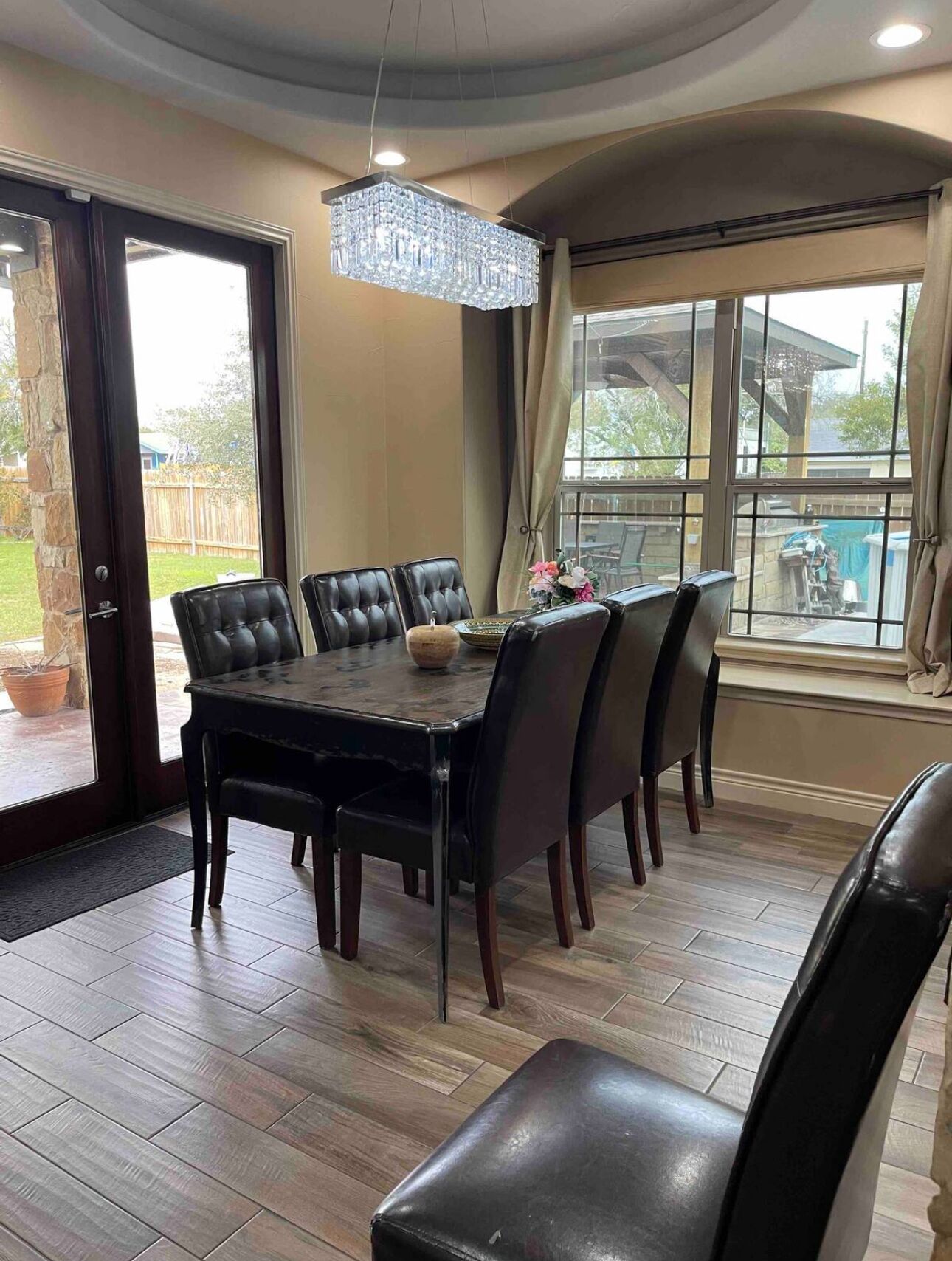 A dining area with a large table and six chairs, a chandelier, and a view through large windows.