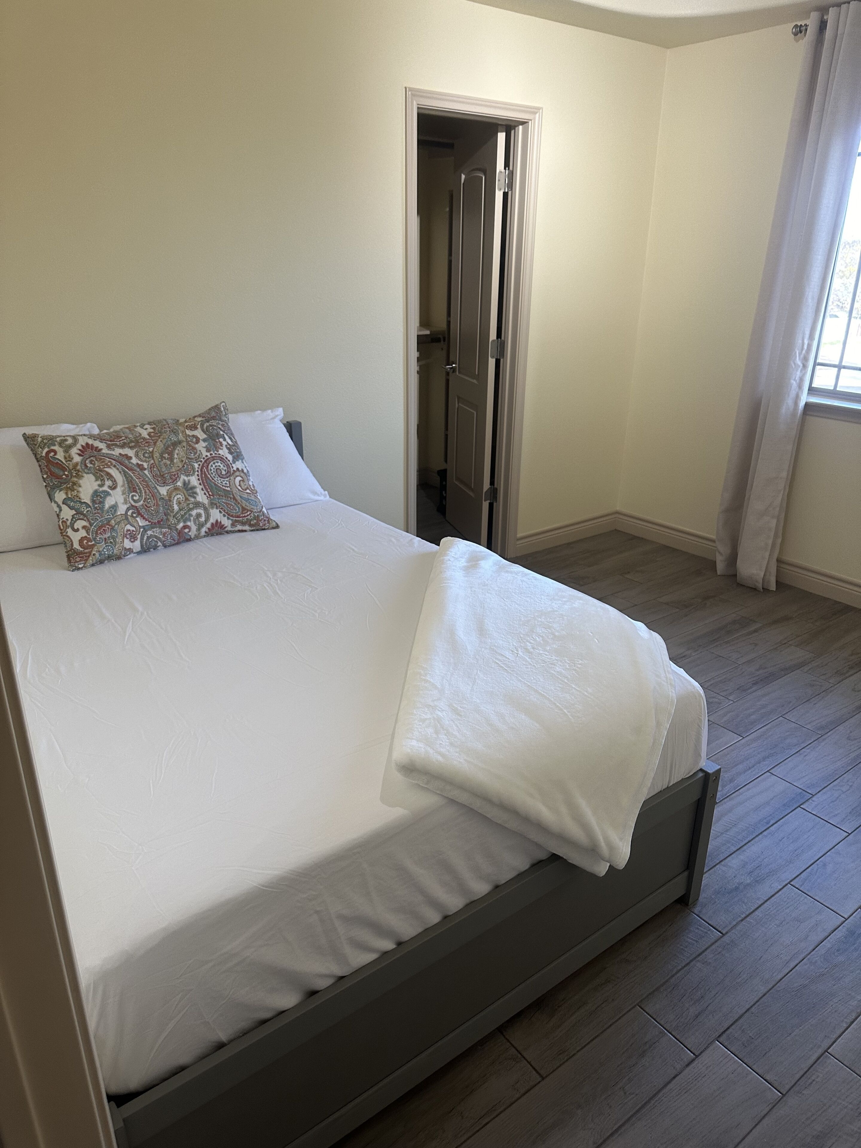 Un lit bien fait, recouvert d’une couvre-lit blanche et agrémenté d’un coussin décoratif, un plancher en bois et une porte donnant sur une autre pièce.
