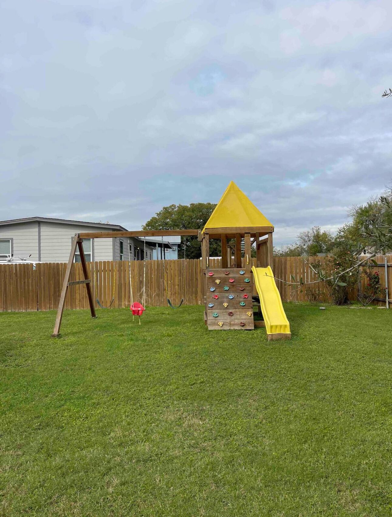 A backyard with a wooden play structure featuring a slide and climbing wall, a swing set, and a fenced area.