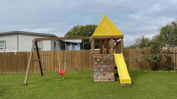 A backyard with a wooden play structure featuring a slide and climbing wall, a swing set, and a fenced area.