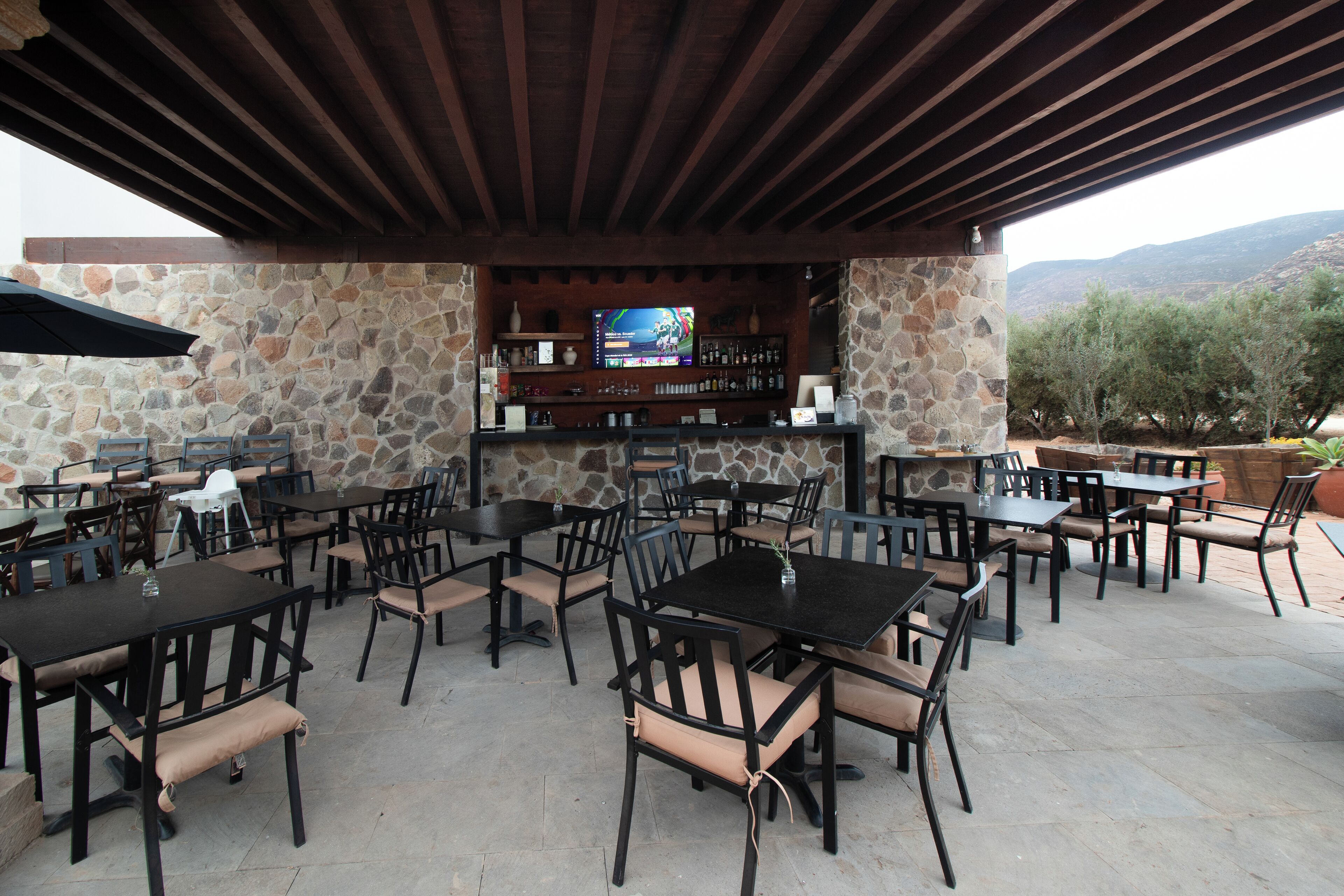 An outdoor patio with stone walls, multiple tables and chairs, a bar area with a television, and a view of the mountains.