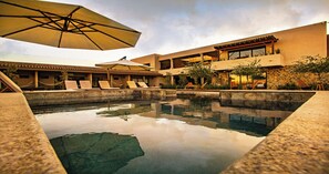 A pool area with lounge chairs and umbrellas, surrounded by buildings with stone walls and large windows. - Hotel Flores de Sal (Valle de Guadalupe)
