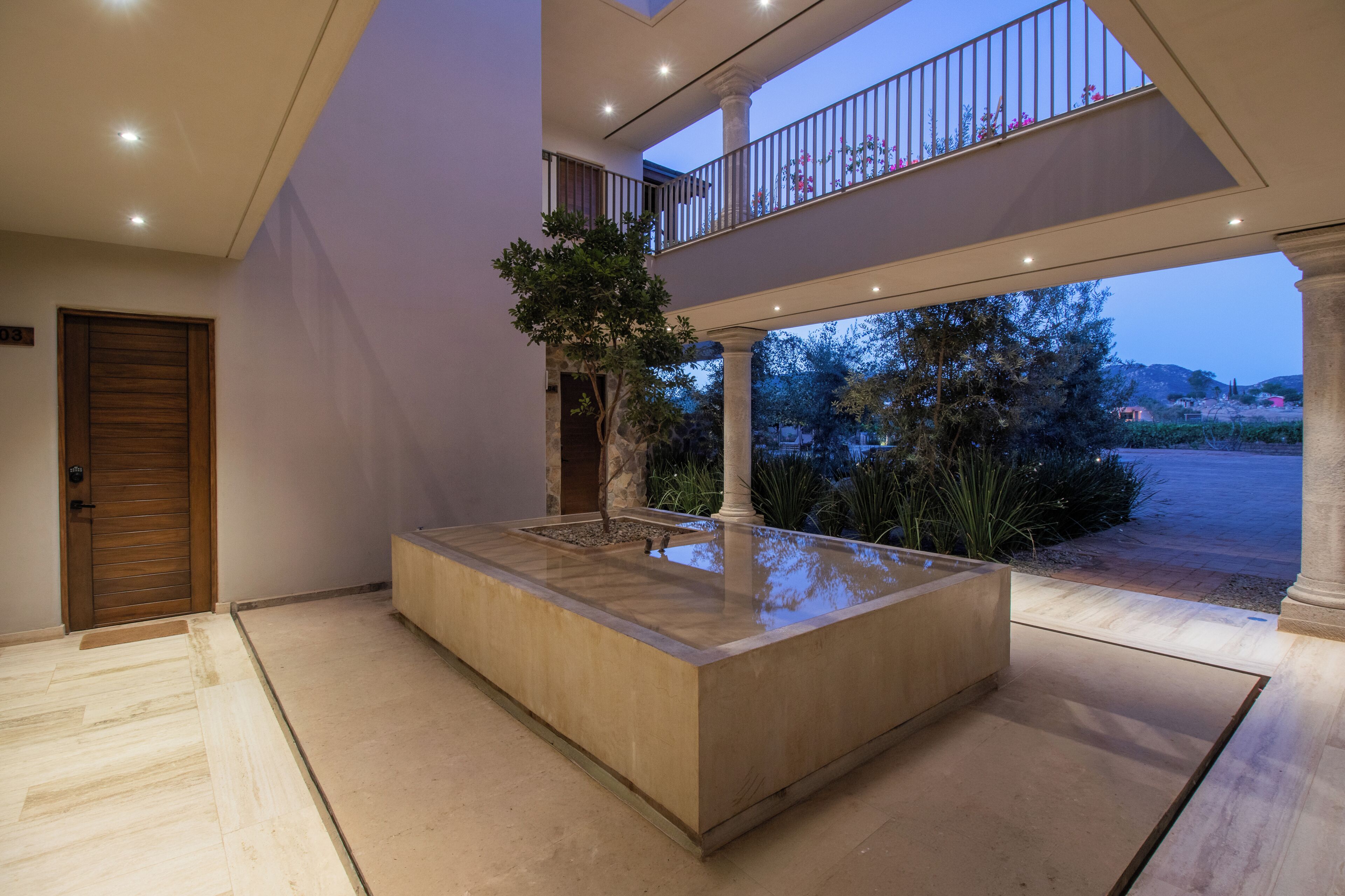 A spacious indoor area with a central water feature, a wooden door, and a balcony overlooking a landscaped garden.