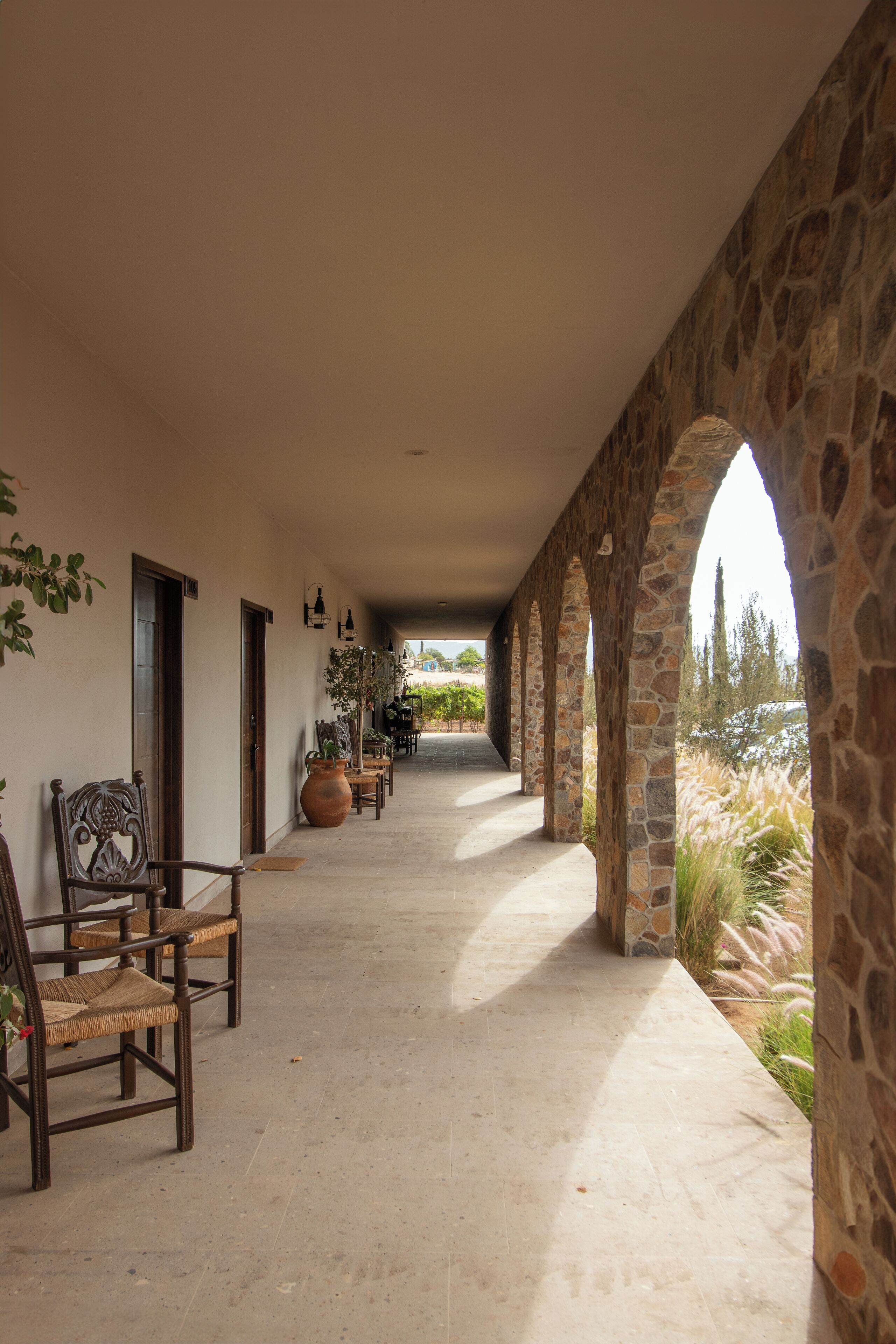 A covered corridor with stone arches, a row of chairs, and potted plants.