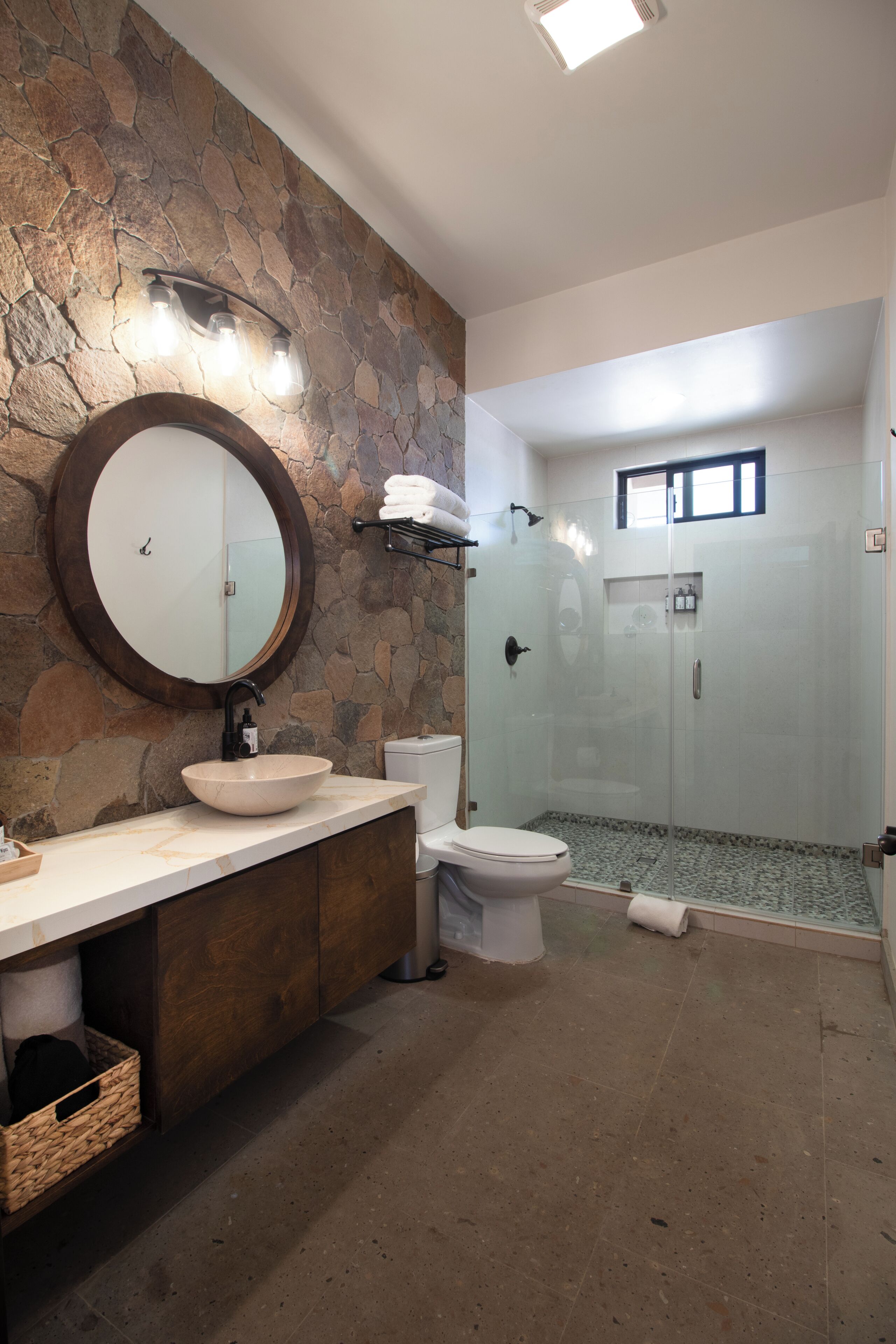 Un baño con pared de piedra, un espejo redondo, lavamanos, inodoro y zona de ducha con mampara de vidrio.