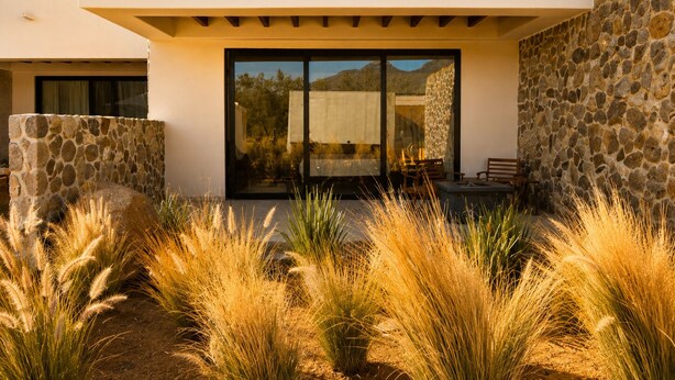 A modern building with a stone facade, large glass doors, and a patio area with tall grasses. - Hotel Flores de Sal (Valle de Guadalupe)