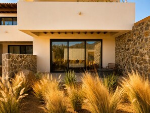 A modern building with a stone facade, large glass doors, and a patio area with tall grasses. - Hotel Flores de Sal (Valle de Guadalupe)
