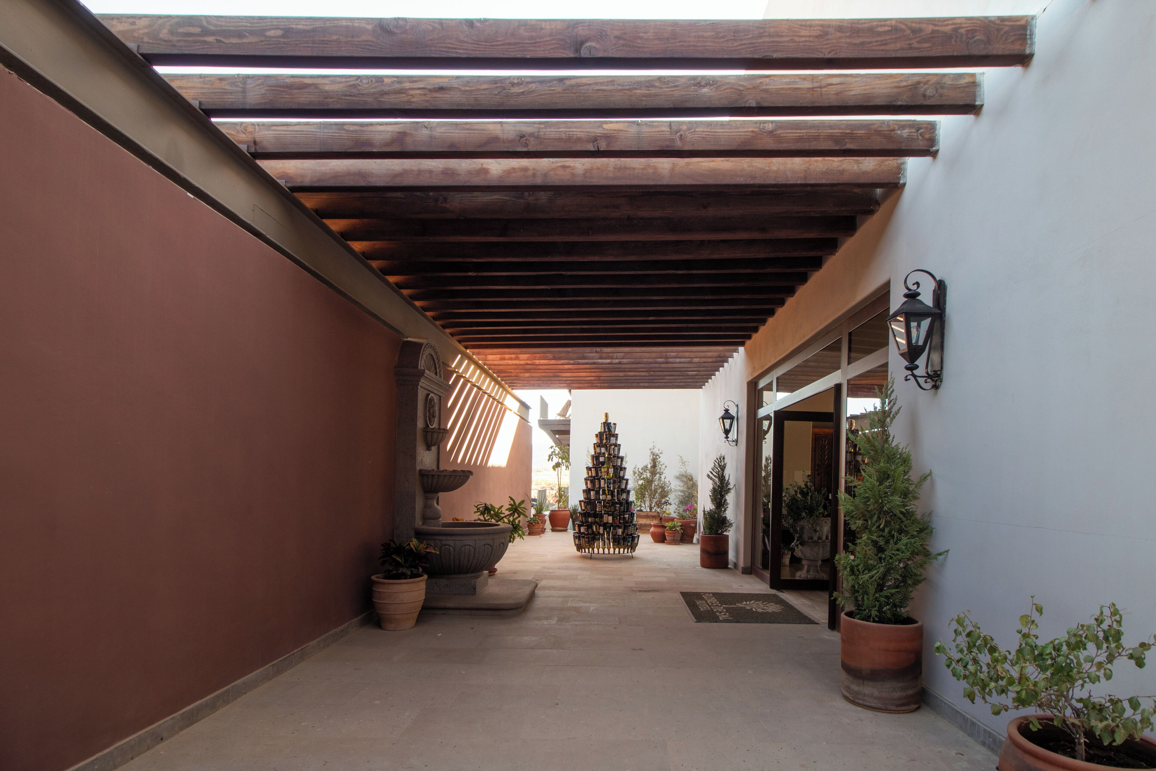 Un espacio al aire libre cubierto con techo de madera, plantas en macetas y una estantería decorativa para vinos.