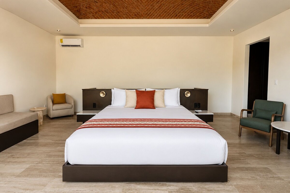 A modern hotel room with a large bed, two nightstands, a sofa, and a chair.