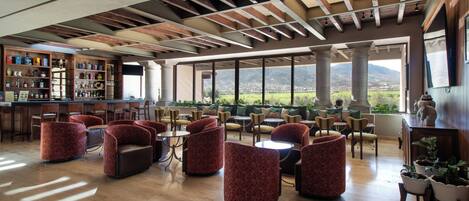 A spacious lounge area with multiple seating options, a bar with a variety of bottles, and large windows offering a view of the landscape.
