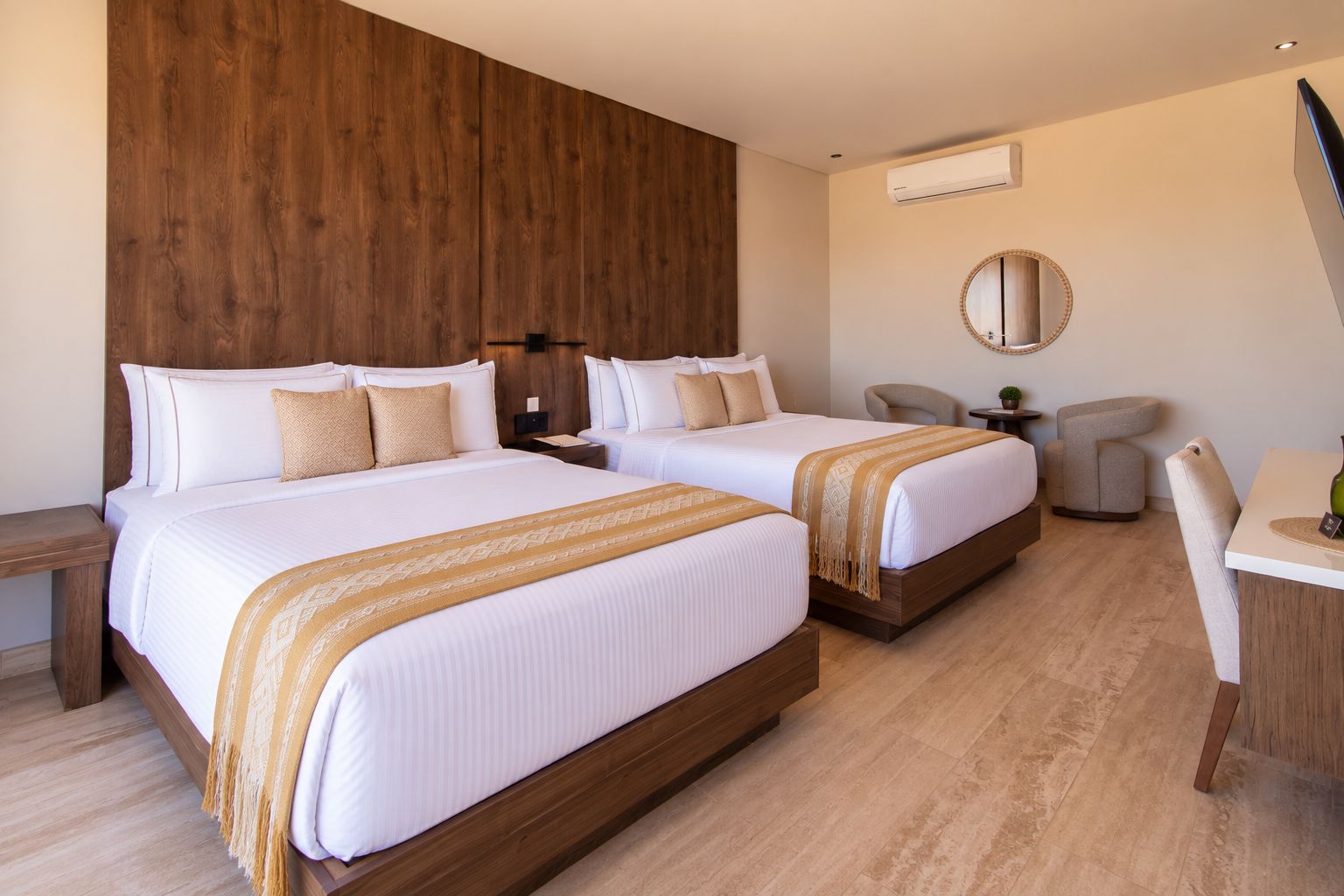 A hotel room with two beds, a desk, a chair, a round mirror, and an air conditioning unit.