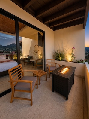 An outdoor patio with a fire table, wooden chairs, a bed, and a view of the landscape.