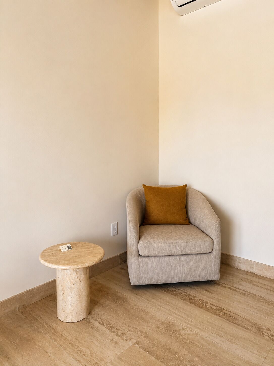 A room with a round wooden table, a beige armchair with a brown cushion, and a light-colored wall.