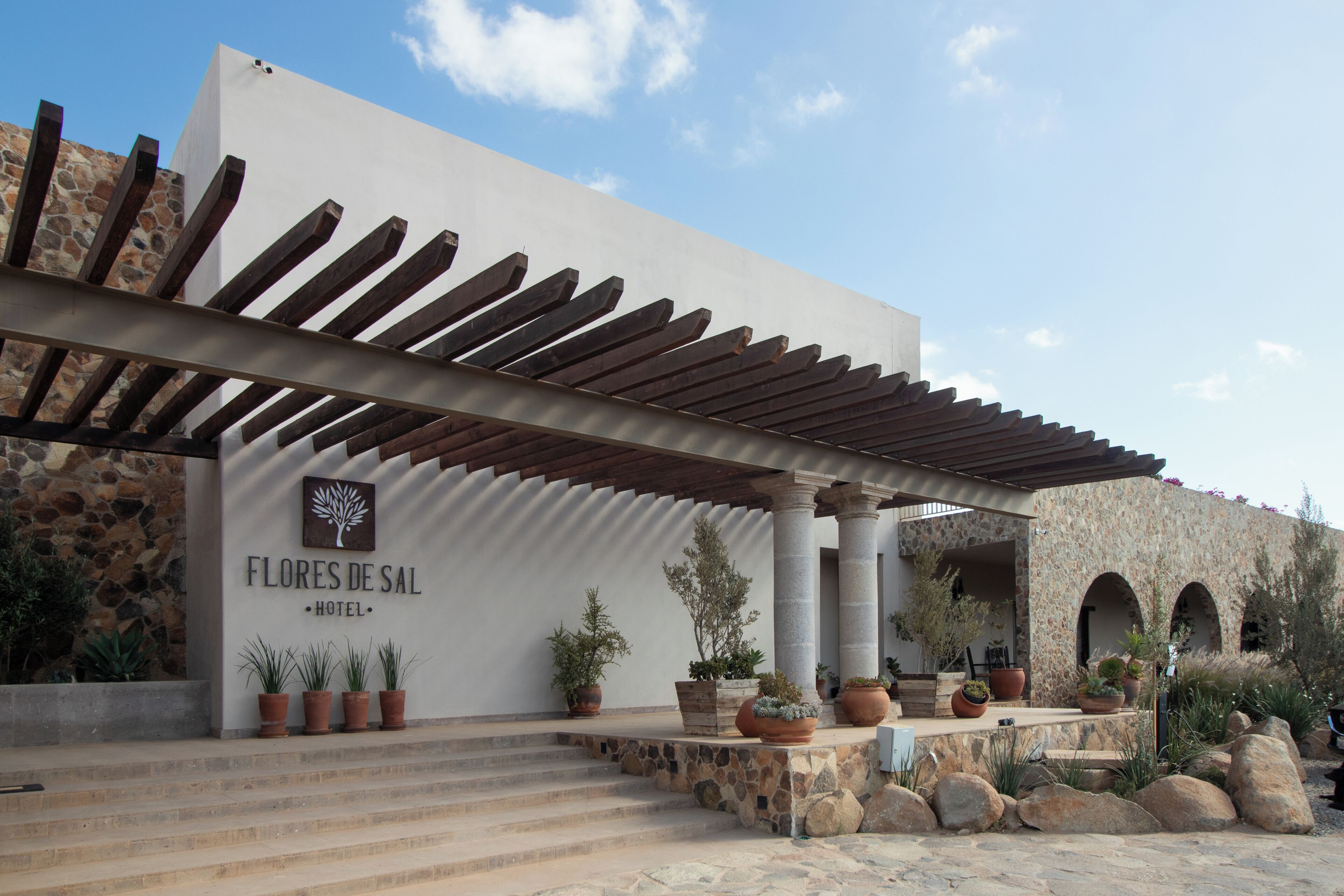 Entrada de un hotel con una pérgola, paredes de piedra y plantas en macetas.