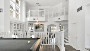 A spacious indoor area with a pool table, a chandelier, and a staircase leading to a balcony.