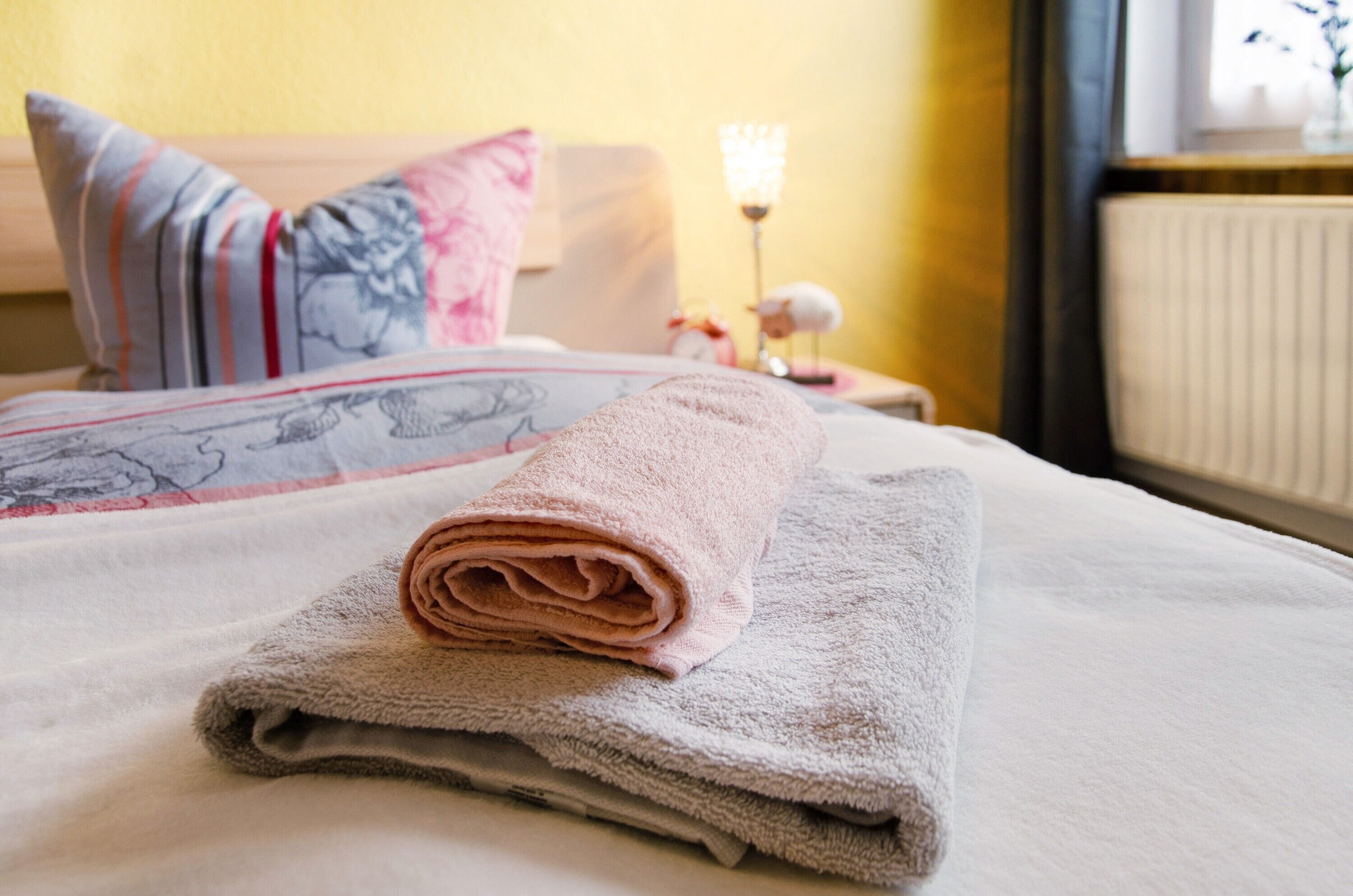 A neatly made bed with a rolled towel, pillows with striped and floral patterns, and a bedside table with a lamp.