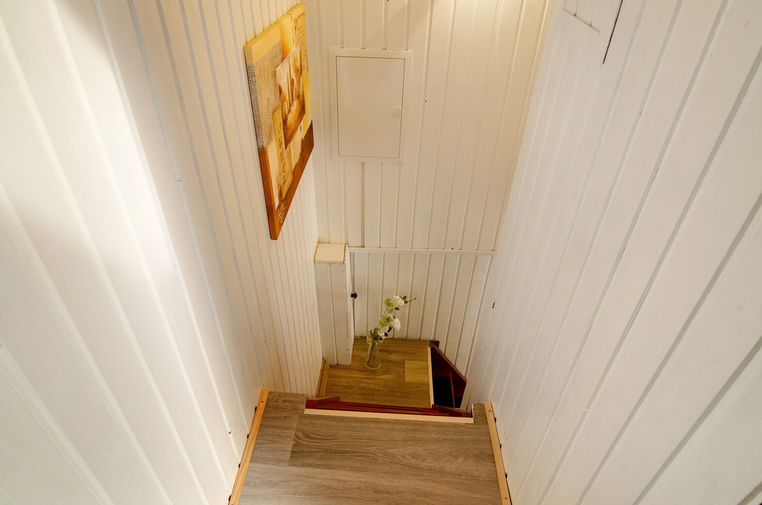 Un couloir étroit aux murs recouverts de lambris en bois, un escalier en bois et un vase rempli de fleurs sur la plateforme.
