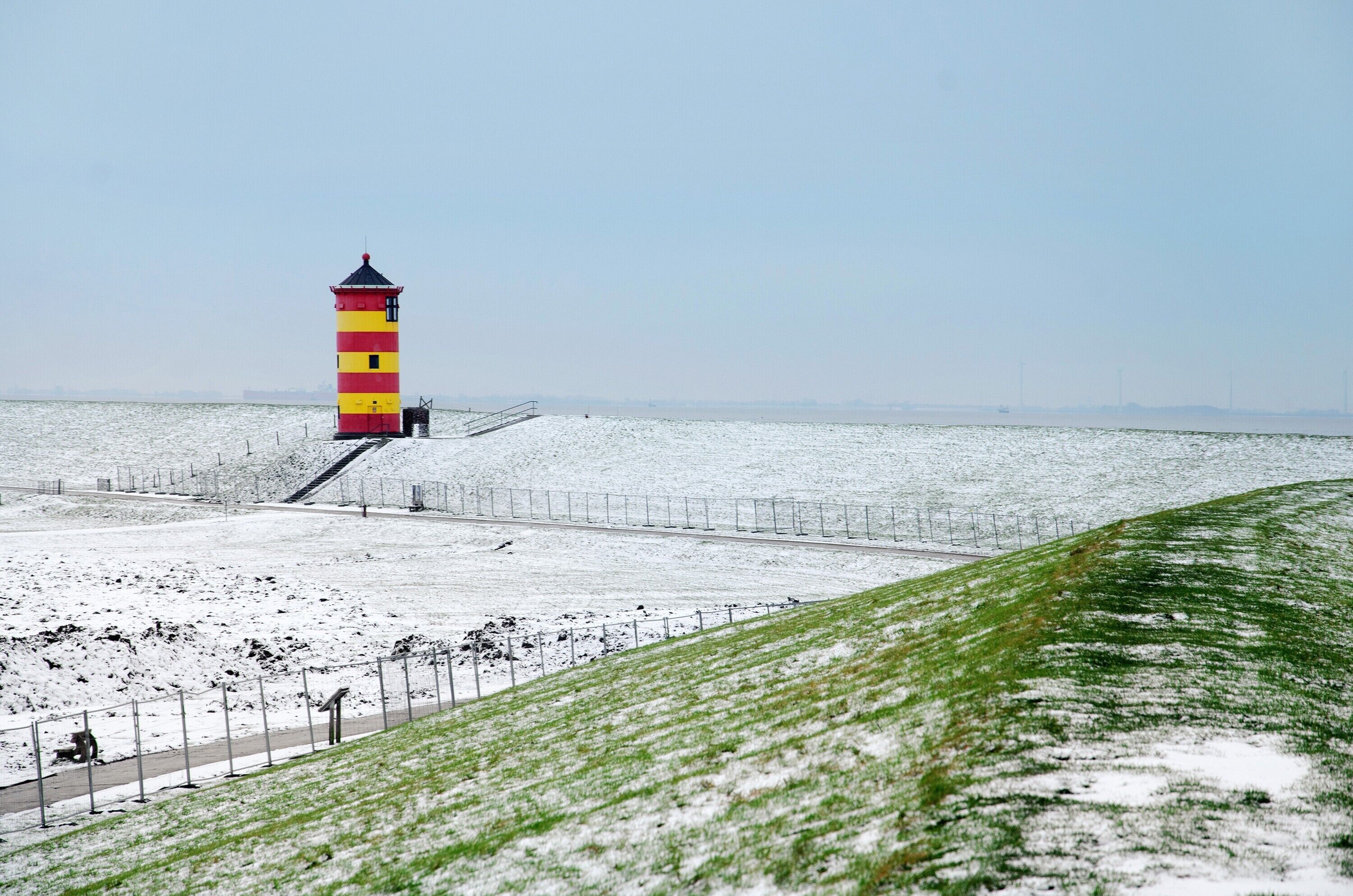 A lighthouse with a red and yellow striped tower, a snow-covered grassy area, and a clear sky.