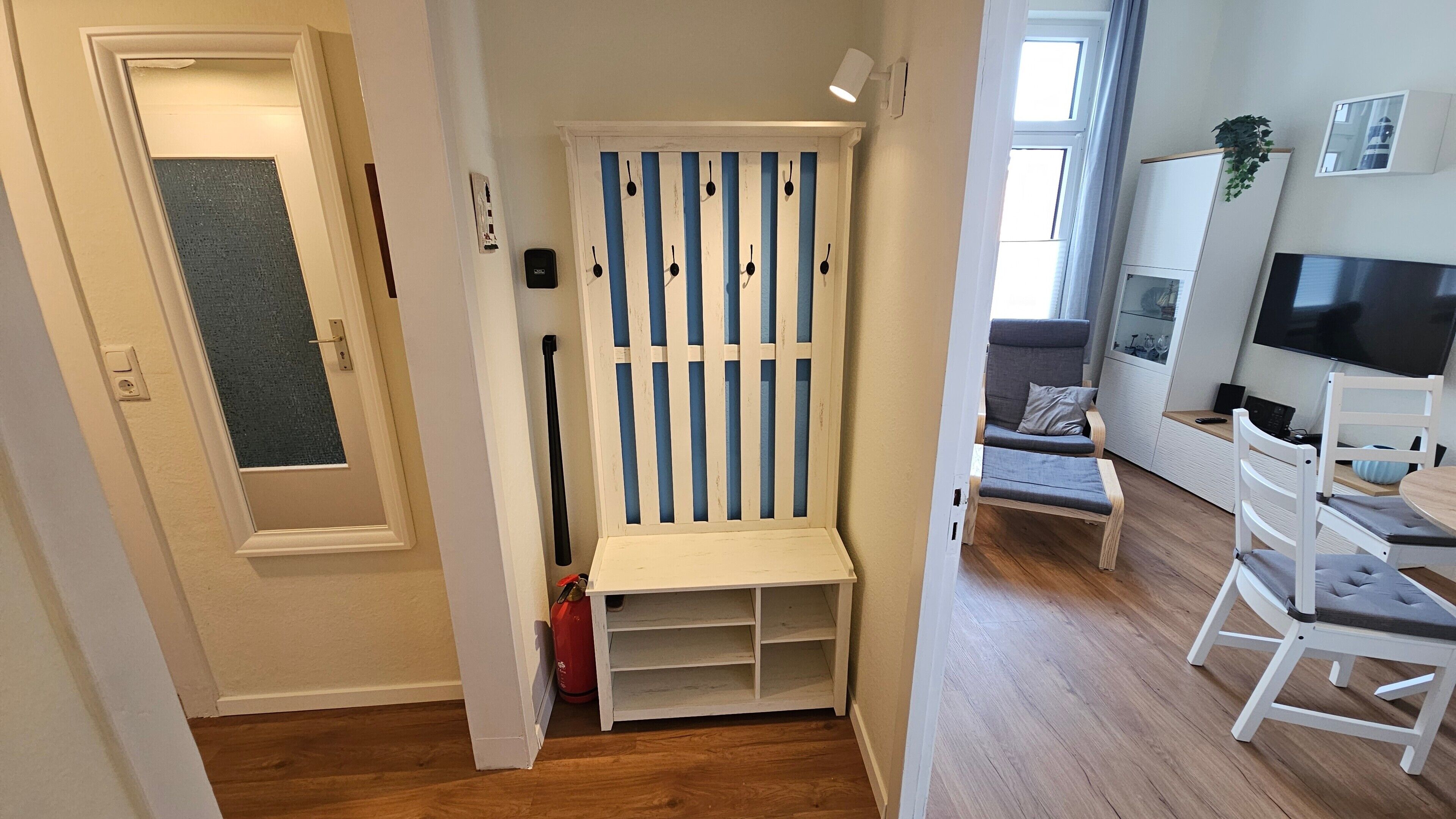 A hallway leading to a room with a white bench, a red fire extinguisher, and a TV in the background.