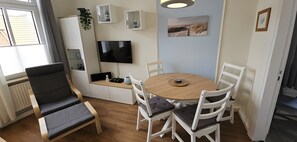 Una habitación con una mesa redonda de madera, sillas blancas, un televisor de pantalla plana, una ventana con persianas y un radiador.