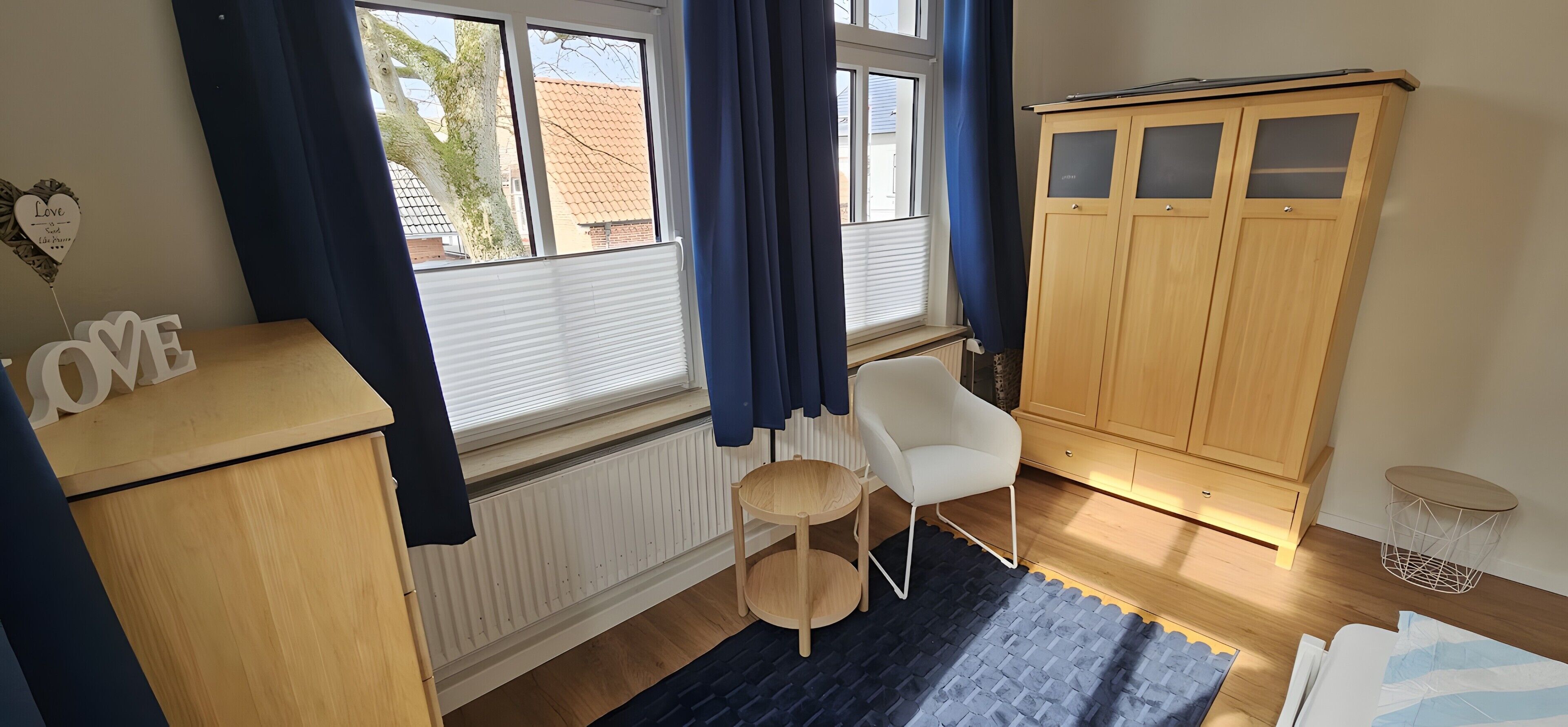 A room with a radiator, a white chair, a wooden cabinet, a wardrobe, and a window with curtains.