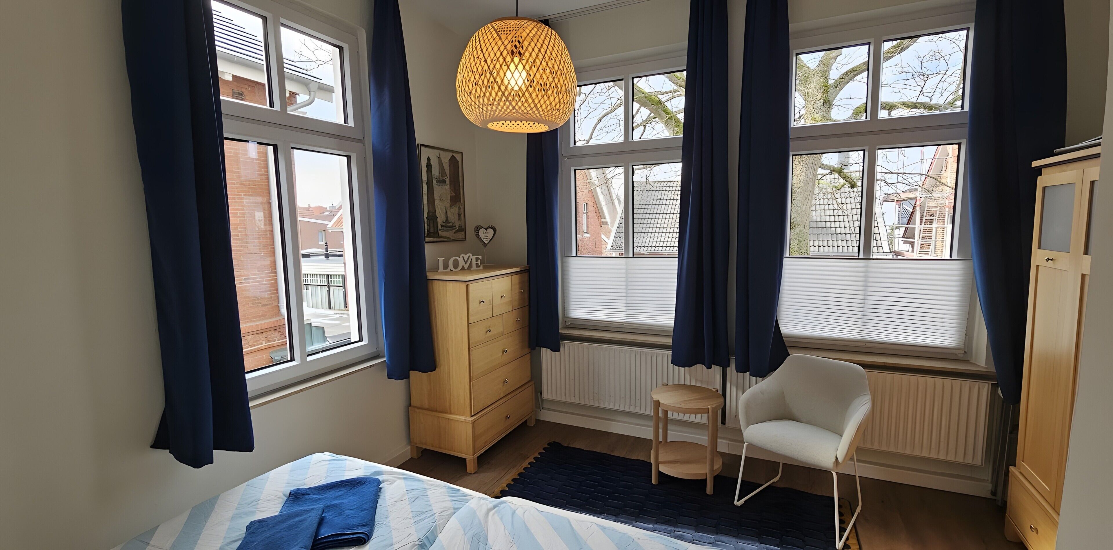 Een slaapkamer met een bed, een kaptafel, een stoel en grote ramen met gordijnen.
