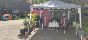 A covered outdoor seating area with a table and chairs, surrounded by potted plants and a play structure in the background.