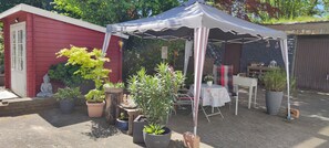 A red building with a white door, a white tent, and various potted plants.