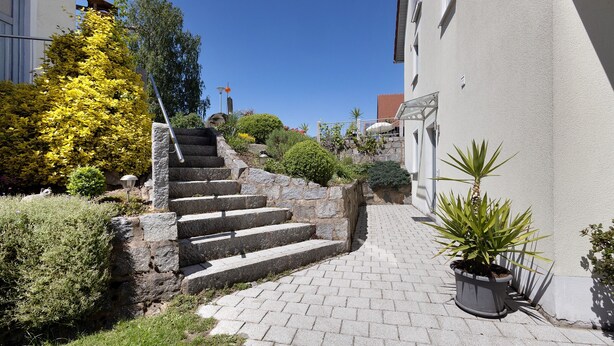 Una escalera de piedra que sube hacia un edificio, rodeada de follaje y un cielo azul despejado.