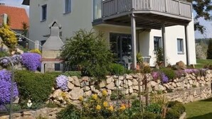 Una casa de dos pisos con un balcón, rodeada por un muro de piedra y diversas plantas.