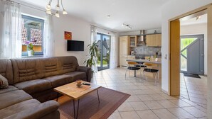 Una sala moderna con un sofá, una mesa de centro y un televisor. Hay una zona de cocina con armarios y una mesa de comedor.