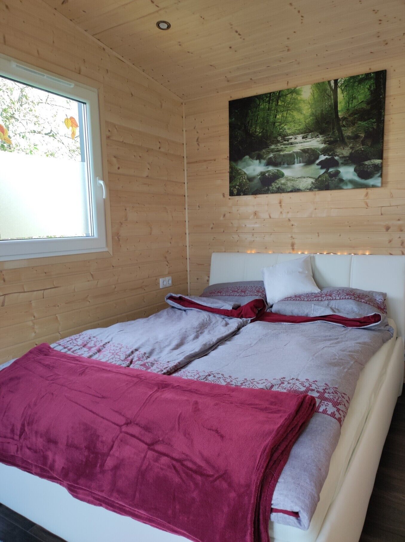 A wooden cabin bedroom with a bed, a window, and a painting of a forest scene.