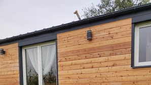 Une cabane en bois avec un banc et des coussins à l’extérieur, une fenêtre et un tuyau d’arrosage sur la pelouse.