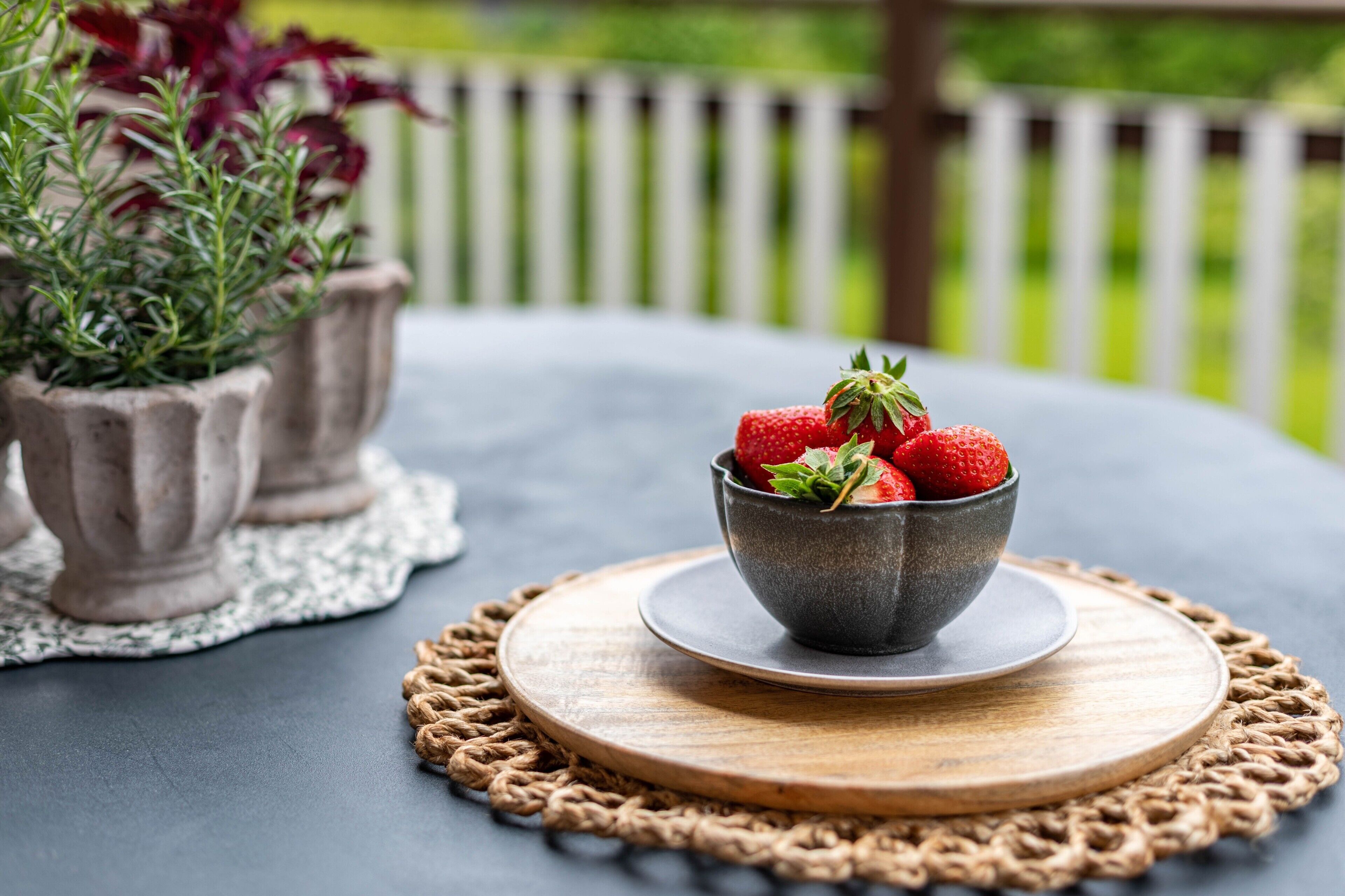 A bowl of strawberries on a table with a placemat and potted plants in the background.