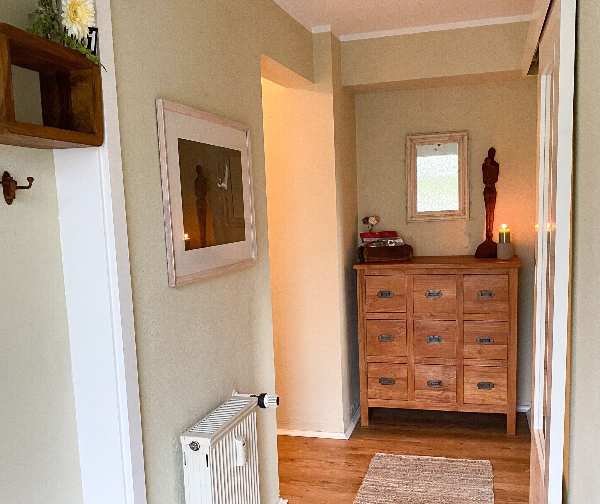 Een hal met een radiator, een houten dressoir met lades, een lijst met een foto aan de muur, en een klein raam.
