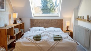 Un dormitorio con tragaluz, una cama con toallas ordenadas, una mesita de noche con lámpara y un estante con objetos decorativos.