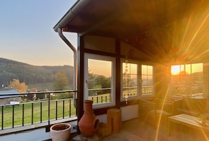 Un balcon donnant sur un lac et des collines, une plante en pot et un tabouret en bois.