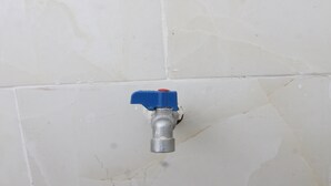 Un robinet mural, fixé au mur, avec une poignée bleue, installé sur un mur carrelé dont on peut voir les joints et qui présente de légères imperfections.