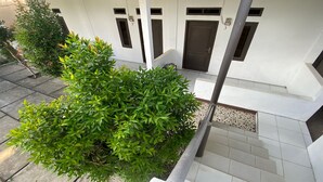 A white building with three doors, numbered 2, 3, and 4, and a small garden area with green shrubs. - OYO Life 93154 White Kos Jenggala (Tanjung)