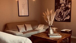 Uma sala de estar aconchegante com um sofá, uma mesa de centro de madeira com um livro e um vaso de capim-do-campo, e uma citação emoldurada na parede.
