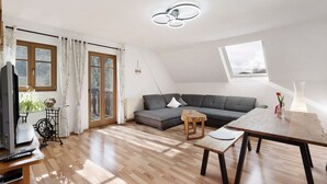 Een modern loft appartement met een groot raam, een houten eettafel, een grijze hoekbank en een televisie op een houten dressoir.