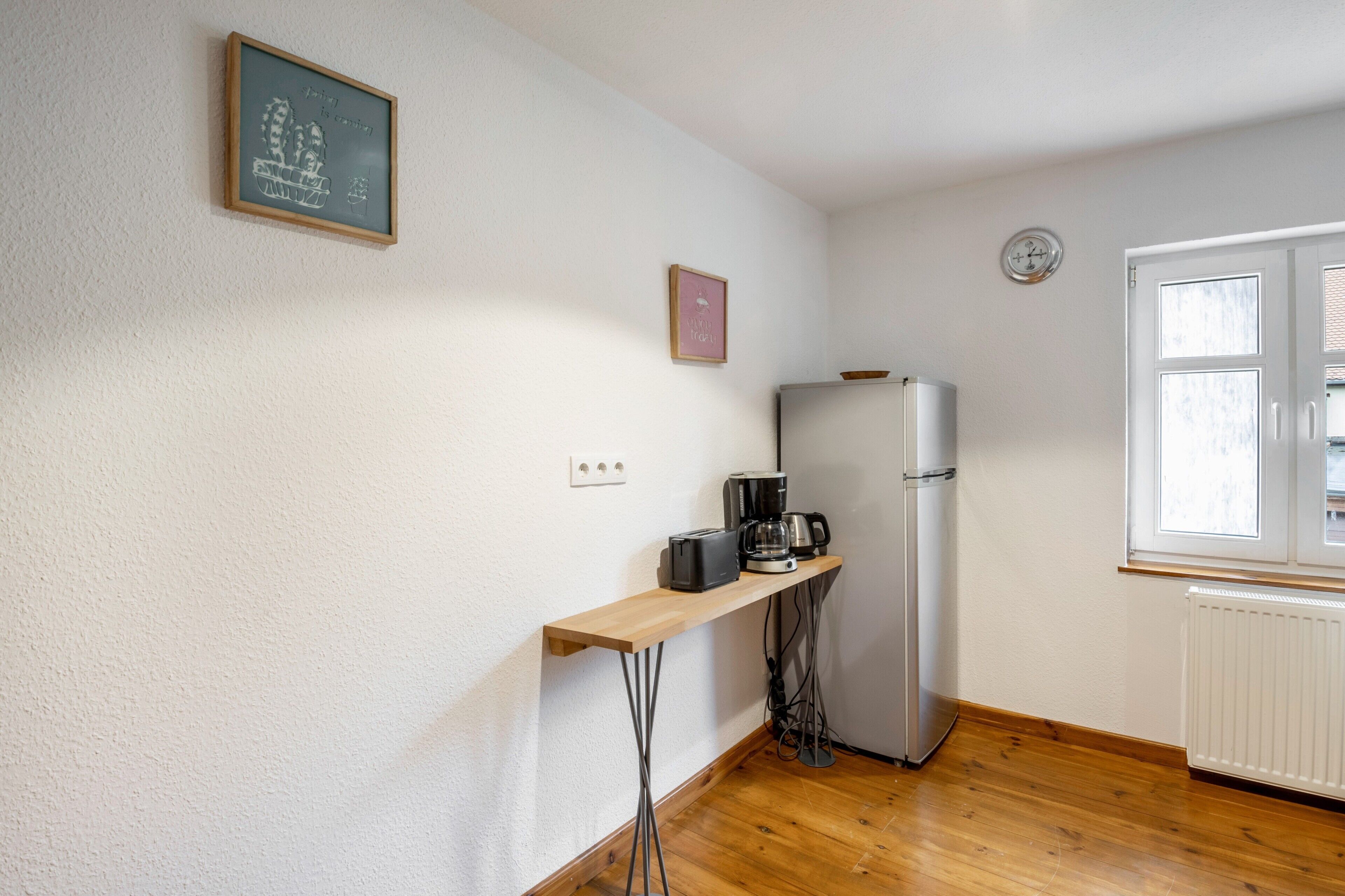 Et lille køkken med et køleskab, kaffemaskine og et vindue.
